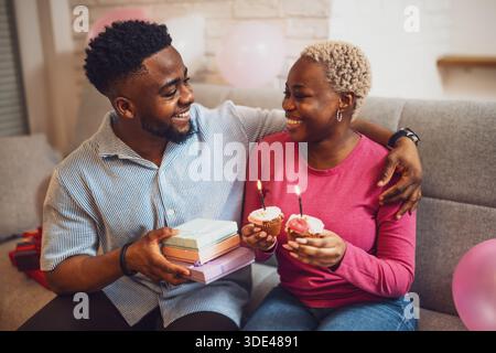 Heureux jeune couple célébrant l'anniversaire de la femme à la maison. Petit ami donne un cadeau à sa petite amie. Banque D'Images