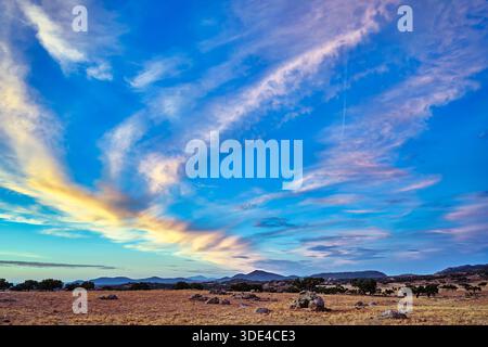 Un ciel de coucher de soleil vibrant affiche des nuages colorés au-dessus d'un paysage rural. La scène présente des champs dorés, des formations rocheuses et des collines lointaines à Extre Banque D'Images