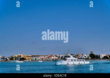 Un bateau blanc se déplace doucement à travers l'eau dans une zone côtière avec des bâtiments visibles le long du rivage. Le ciel bleu vif améliore le paysage. Banque D'Images
