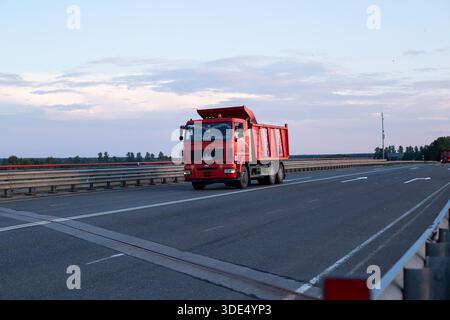 Minsk, Biélorussie 20.06.23 : un gros camion à benne rouge traverse un pont autoroutier à plusieurs voies au crépuscule. Carrosserie peinte rouge vif, caisse emp Banque D'Images