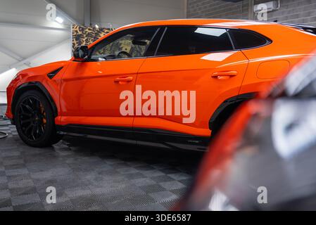 Une vue latérale rapprochée montre une Lamborghini Urus se orange garée dans un atelier moderne, avec peinture brillante, roues noires, étriers orange, floo à carreaux Banque D'Images