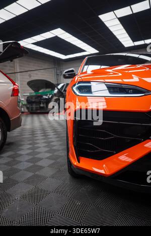 Fermer vue de trois quarts montre Lamborghini Urus se orange dans un atelier, phare LED, pare-chocs sculpté, calandre noire brillante, voitures à proximité avec capot an Banque D'Images