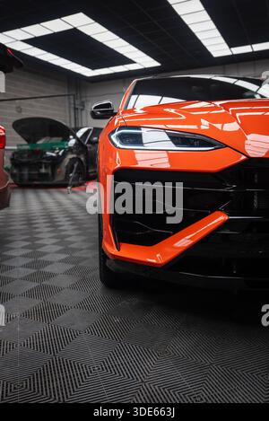 Une vue rapprochée montre une Lamborghini Urus se avant orange, un phare LED, une grande calandre noire et des ailettes pointues dans un atelier avec des voitures en service et fl Banque D'Images