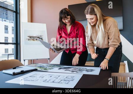 Deux jeunes femmes discutent de plans architecturaux à une table de bureau moderne, partageant des idées. Banque D'Images