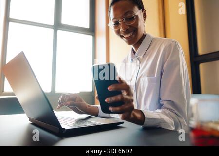 Une jeune femme africaine confiante travaille avec un ordinateur portable et un téléphone dans un cadre de bureau moderne. Productivité, communication et technologie dans une profession Banque D'Images
