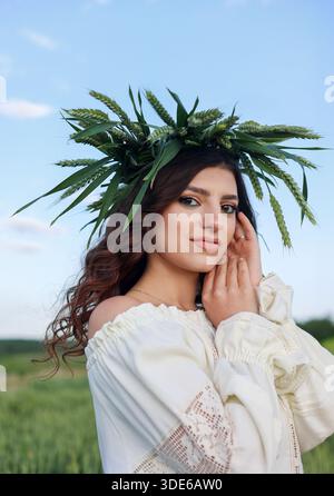 Belle jeune femme en robe brodée traditionnelle et couronne de blé vert regardant la caméra dans un champ de seigle luxuriant. Banque D'Images