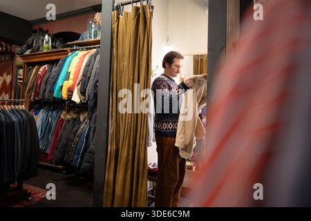 Homme élégant client magasinant des vêtements vintage dans le magasin de seconde main, essayer la veste dans la cabine d'essayage. Banque D'Images