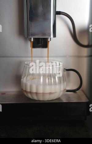 une machine à café distribuant un espresso riche dans une tasse en verre transparent remplie de lait crémeux, créant une apparence en couches, sertie sur une surface métallique Banque D'Images