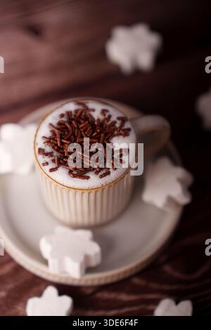 ambiance chaleureuse et accueillante avec une tasse de cappuccino mousseux garni de chocolat, entourée de guimauves en forme de flocon de neige sur une céramique Banque D'Images