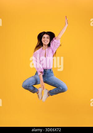 Joyeuse femme arménienne sautant et touchant les jambes avec la main, fond jaune de studio, pleine photo Banque D'Images