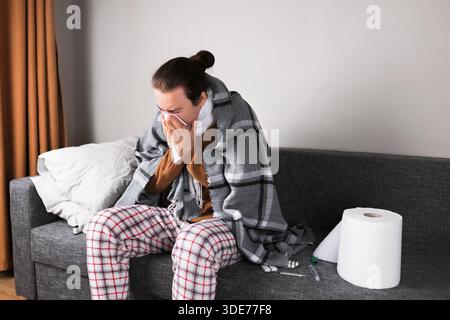 Homme malade dans la couverture soufflant le nez dans le tissu sur le canapé avec thermomètre et pilules à la maison. Photo de haute qualité Banque D'Images
