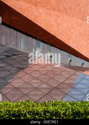 Vue abstraite d'un coin de bâtiment présentant un mur rouge texturé, un pavé géométrique gris carrelé et une bordure de haie verte. L'image se concentre sur des lignes Banque D'Images