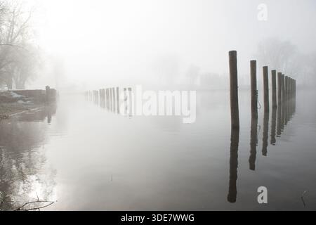Le brouillard entoure un lac, obscurcissant la rive lointaine. Des poteaux en bois se dressent dans l'eau, vestiges d'un quai, créant une scène sereine et minimaliste. Banque D'Images