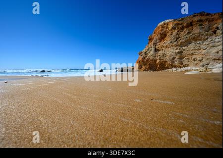 Le sable doré rencontre la mer turquoise sur une plage isolée. Les vagues roulent doucement, créant une scène paisible sous un ciel bleu clair, parfait pour se détendre Banque D'Images