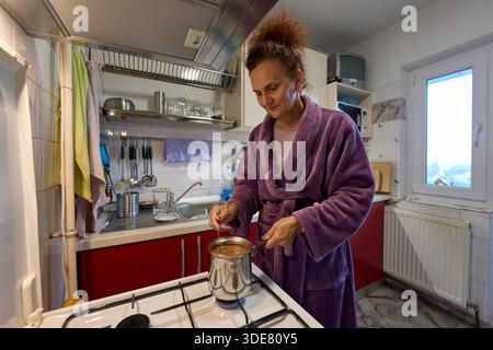 Femme en peignoir violet remue le pot de café turc sur la cuisinière à gaz dans la cuisine pendant la routine du matin Banque D'Images
