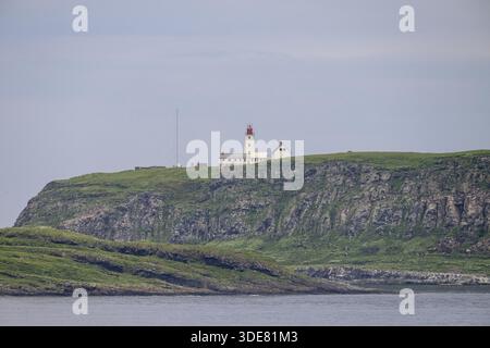 Phare blanc avec pointe rouge et antennes s'élève au-dessus des rochers abrupts sur l'île d'oiseaux de Hornoya, entouré de collines herbeuses sous un ciel clair, Vardo Banque D'Images