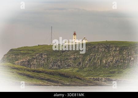 Phare blanc avec pointe rouge et antennes s'élève au-dessus des rochers abrupts sur l'île d'oiseaux de Hornoya, entouré de collines herbeuses sous un ciel clair Un cadre Banque D'Images