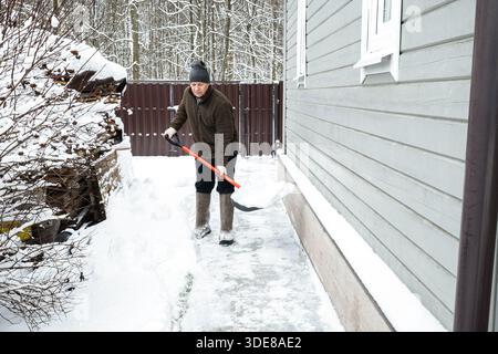 Homme âgé dégageant la neige avec une pelle près de la maison dans la cour d'hiver. Travail saisonnier en plein air, style de vie rural et routine hivernale avec déneigement. Banque D'Images