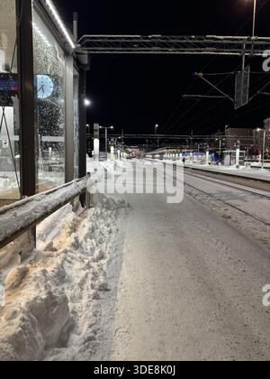 Quai de train enneigé la nuit dans une ville en hiver, avec des voies vides et une infrastructure de gare éclairée. Banque D'Images