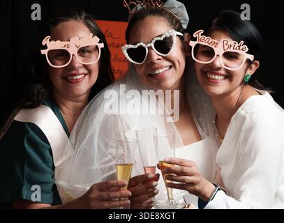 Trois femmes portent des lunettes de soleil et tiennent des verres de champagne. L'une des femmes porte une robe de mariée Banque D'Images