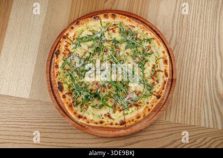 Une pizza ronde à croûte fine est fraîchement cuite et présentée sur un plateau en bois. Il se compose d'un mélange de fromage fondu, de roquette fraîche et d'une touche de p Banque D'Images