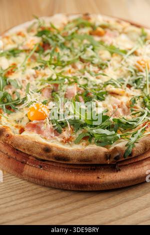 Une pizza cuite au feu de bois présente une fine croûte recouverte de roquette, de jambon, de fromage et d'oeuf sur une planche de bois. Parfait pour partager ou profiter seul. Banque D'Images
