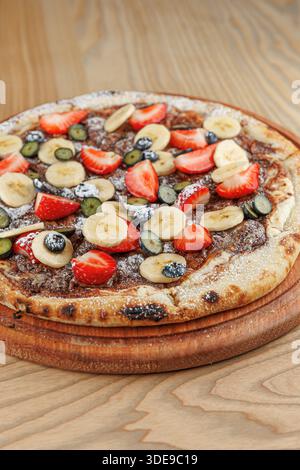 Une pizza dessert à la croûte fine est garnie de fraises fraîches, de bananes et de bleuets. La pizza est placée sur un plateau de service en bois, montrant Banque D'Images