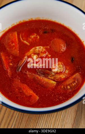 Un bol de soupe rouge rempli de fruits de mer, de tomates et d'autres légumes est placé sur une table en bois. La soupe semble copieux et savoureux, prêt à en Banque D'Images