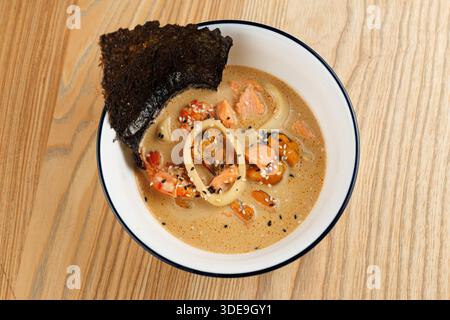 Un bol de ramen rempli de bouillon, de crevettes, de saumon et d'algues se trouve sur une table en bois. Le plat met en valeur des saveurs riches et des ingrédients de fruits de mer frais. Banque D'Images