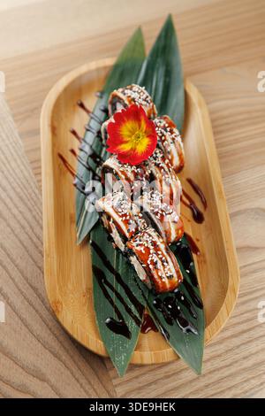 Les rouleaux de sushi sont agencés habilement sur une assiette de bambou avec des feuilles et une fleur vibrante. Un filet de sauce ajoute du contraste à la présentation. Banque D'Images