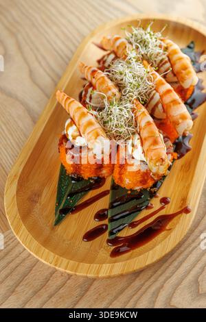 Divers rouleaux de sushi reposent sur un plateau en bois. Chaque rouleau est recouvert de crevettes et décoré de germes. Un filet de sauce ajoute de la saveur au plat. Banque D'Images