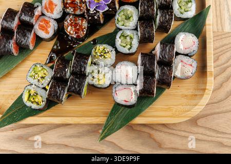 Des rouleaux de sushi avec diverses garnitures sont disposés soigneusement sur un plateau en bambou avec des feuilles vertes fraîches. Cet affichage met en évidence une variété de couleurs et de textures Banque D'Images