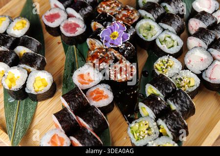 Divers rouleaux de sushi sont disposés sur un plateau en bois avec des feuilles vertes. Les rouleaux comprennent diverses garnitures et garnitures, mettant en valeur la cuisine japonaise Banque D'Images