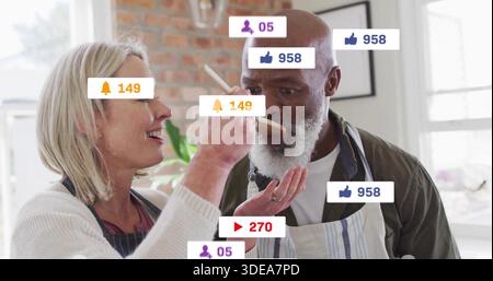 Offrant femme portant tabliers tenant cuillère en bois nourrissant homme barbu à la cuisine de la maison, avec des badges Banque D'Images