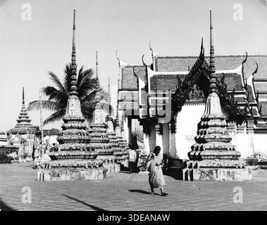 Blick auf die kunstvollen Chedis und Tempelgebäude des Wat Pho à Bangkok. Eine person geht über den gepflasterten Innenhof der Anlage. Thaïlande, 1960er. Banque D'Images