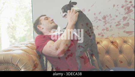 Tenant l'homme en chemise rouge soulevant le chien noir et blanc sur le canapé touffeté à la maison, avec fenêtre rétro-éclairée Banque D'Images