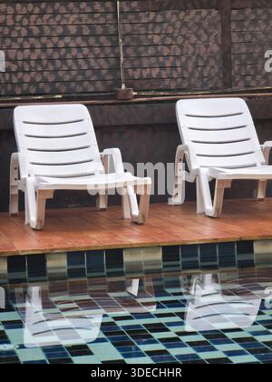 Atmosphère relaxante au bord de la piscine avec deux chaises longues en plastique blanc vides reflétant dans l'eau bleue calme de la piscine en attente des vacances d'été gue Banque D'Images