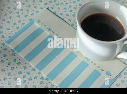 Une tasse de café noir confortable se tient sur un fond floral, et une lettre se trouve à côté Banque D'Images
