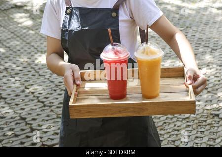 Plan recadré de serveuse portant un plateau en bois avec smoothie aux fruits rafraîchissants froids dans une tasse en plastique servant le client avec une émotion accueillante à l'extérieur Banque D'Images