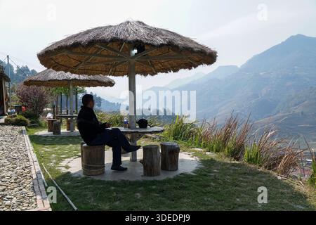 L'homme bénéficie d'une vue panoramique sur les montagnes brumeuses et les champs en terrasses d'un point de vue serein sous un parapluie de chaume. Banque D'Images