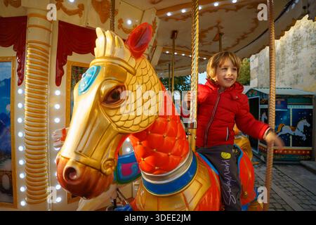 Enfant souriant en veste rouge chevauchant un cheval de carrousel coloré dans un parc d'attractions en plein air pendant la journée Banque D'Images