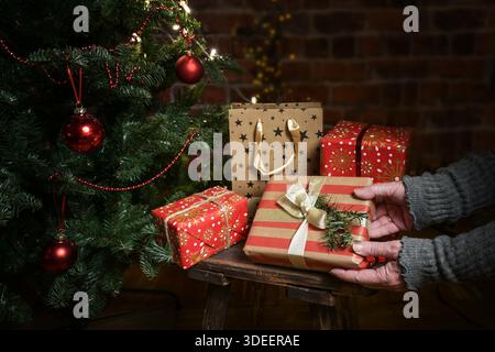 Mains d'une femme plus âgée plaçant des cadeaux emballés sous un arbre de Noël décoré contre un mur de briques sombres, concept pour donner avec amour et espace de copie de don, foyer sélectionné, profondeur de champ étroite Banque D'Images