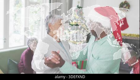 Couple senior dansant tenant la main dans le salon, arbre visible, chapeau de Père Noël et cardigan gris Banque D'Images
