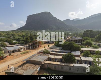 Vue aérienne d'une montagne accidentée se profile au-dessus d'un village de bâtiments aux toits d'étain, avec des arbres rouges vibrants ponctuant les tons terreux, VOI, Taita-Taveta Banque D'Images