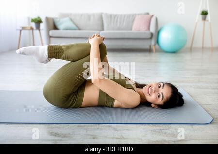 Bonne tenue femme indienne étirant son bas du dos sur le tapis de yoga, embrassant ses genoux, faisant des sports domestiques à l'intérieur Banque D'Images