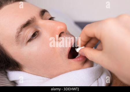 Gros plan d'un jeune homme malade avec une écharpe ouvrant la bouche pour prendre une pilule blanche de la main d'une autre personne. Photo de haute qualité Banque D'Images