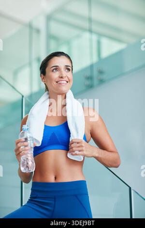 Fitness, pensée et femme heureuse avec de l'eau dans la salle de gym, réflexion d'entraînement ou hydratation post-entraînement. Souriez, terminez l'exercice ou la personne réfléchie avec Banque D'Images