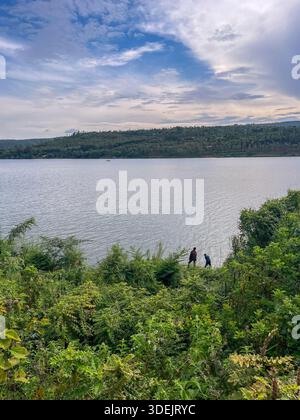 Vue sur un lac calme depuis une rive verdoyante avec deux personnes debout au bord de l'eau au Rwanda. Banque D'Images