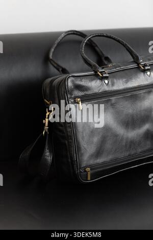 Une mallette professionnelle en cuir noir avec deux poignées et lanière réglable se trouve sur un élégant canapé en cuir noir dans un cadre de studio minimaliste Banque D'Images