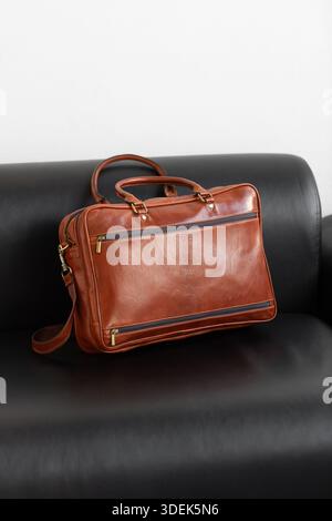 Une mallette professionnelle en cuir cognac avec deux poignées et lanière réglable se trouve sur un élégant canapé en cuir noir dans un cadre de studio minimaliste Banque D'Images